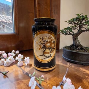 Puede incluir: Un jarrón negro con detalles dorados y un diseño floral decorativo. El jarrón tiene forma cilíndrica y está sobre una superficie de madera. También se ven un bonsái y ramas florecidas.