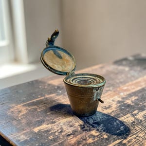 May include: A vintage brass inkwell with a hinged lid, open to show the inside. The inkwell has a small handle and sits on a wooden surface. The brass has a weathered, aged look, indicating its age and history.