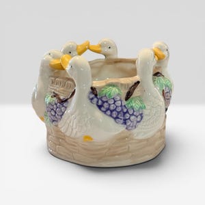 May include: A ceramic planter featuring a circular basket-weave base in a light tan color. The exterior is decorated with several white ducks with yellow beaks, interspersed with clusters of purple grapes and green leaves around the rim.