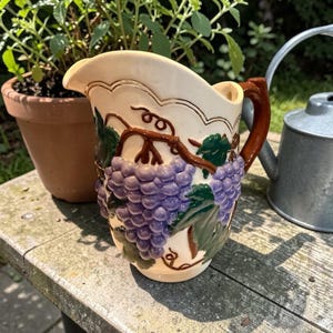 May include: Cream-colored ceramic pitcher with a brown handle and spout. The pitcher is decorated with a cluster of purple grapes, green leaves, and brown vines. The pitcher is sitting on a wooden surface.