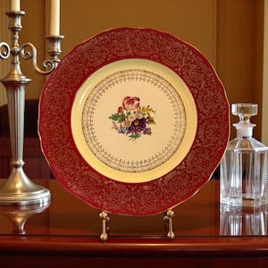 May include: A decorative plate with a red and gold ornate border and a cream-colored center. The center features a floral bouquet. The plate is displayed on a gold stand, with a glass decanter visible in the background.
