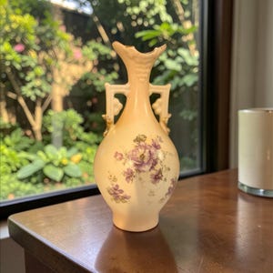 May include: A cream-colored porcelain vase with a bulbous body and a narrow neck featuring two ornate handles. The surface is decorated with a delicate floral pattern of purple flowers and green leaves, accented with gold trim along the rim and handles.