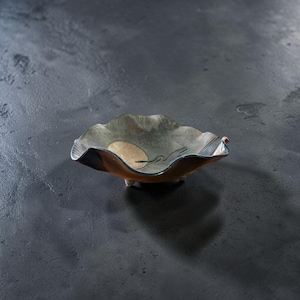Op de afbeelding: Een ondiepe, op voetjes staande keramische schaal met een golvende, organische rand. De binnenzijde heeft een metallic, iriserende afwerking in zilver- en bronskleuren, met een glad, beige cirkelvormig accent en een donkere, abstracte lijntekening.