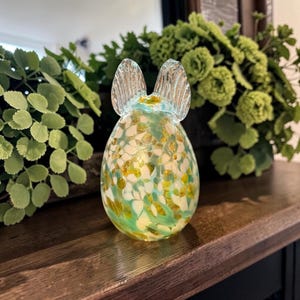 May include: A decorative glass bunny figurine with a speckled pattern of white, yellow, and green. The bunny has clear, textured ears and a light blue accent at the top. The figurine is displayed on a wooden surface.