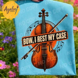 Könnte beinhalten: Ein hellblaues T-Shirt mit einer Grafik einer braunen Holzgeige und einem Bogen. Der Text "BOW, I REST MY CASE" ist in Schwarz auf einem orangefarbenen rechteckigen Hintergrund in der Mitte gedruckt. Noten umgeben das Instrument.