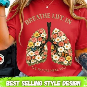 以下が含まれることがあります： 「BREATHE LIFE IN」と「LET NATURE HEAL YOU」の文字が入った赤いTシャツ。蝶と花の肺のデザインが特徴です。Tシャツの下部には「BEST SELLING STYLE DESIGN」の文字が入っています。