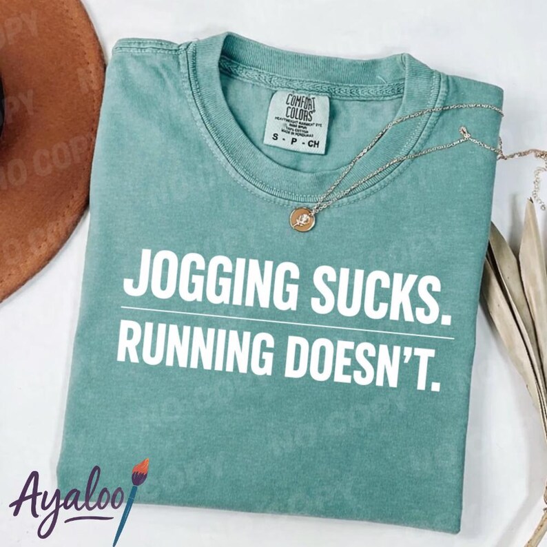 Puede incluir: Una camiseta Comfort Colors de color verde azulado con el texto "JOGGING SUCKS. RUNNING DOESN'T." en blanco. Un collar con colgante dorado descansa sobre la camisa. La camisa es talla S-CH.