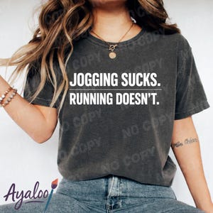 Könnte beinhalten: Dunkelgraues T-Shirt mit dem weißen Text "JOGGING SUCKS. RUNNING DOESN'T.". Das Shirt ist ein lässiger Stil, geeignet für den Alltag. Das Bild zeigt auch eine Person, die das Shirt trägt.