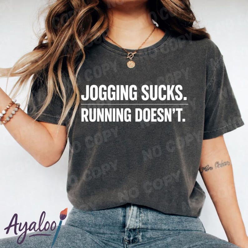 Puede incluir: Camiseta gris oscuro con el texto blanco "JOGGING SUCKS. RUNNING DOESN'T.". La camiseta es de estilo casual, adecuada para el uso diario. La imagen tambi&eacute;n muestra a una persona que lleva la camiseta.