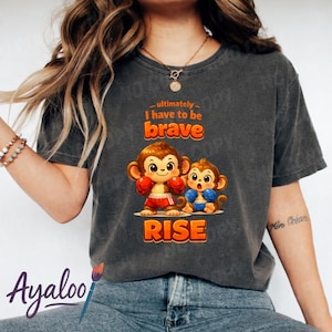 Op de afbeelding: Donkergrijs T-shirt met twee cartoonapen. Eén aap draagt rode bokshandschoenen en een korte broek, de andere een blauwe korte broek. De tekst "ultimately I have to be brave RISE" is gedrukt in oranje en wit.