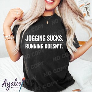 Puede incluir: Camiseta negra con el texto blanco "JOGGING SUCKS. RUNNING DOESN'T.". La camiseta es de estilo casual, adecuada para el uso diario. La camiseta es de color oscuro.