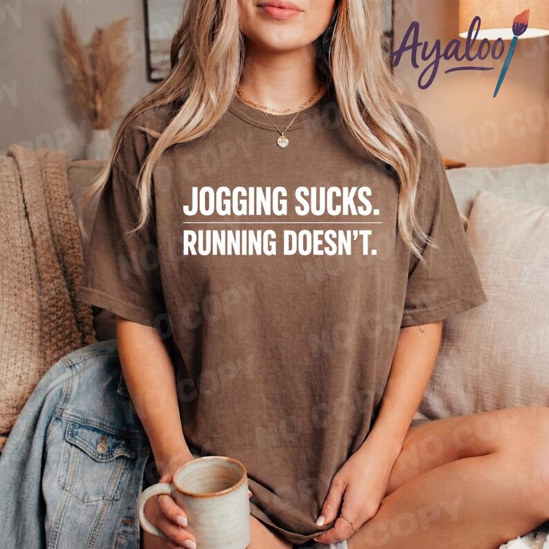 Puede incluir: Una camiseta marr&oacute;n con el texto "JOGGING SUCKS. RUNNING DOESN'T." en blanco. La camiseta es de estilo casual, adecuada para el uso diario. La imagen muestra la camiseta en una persona.