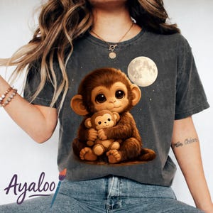 Peut inclure: T-shirt gris foncé avec un singe de dessin animé tenant un singe en peluche. Une pleine lune et des étoiles sont en arrière-plan. Le mot "Ayaloo" est imprimé en cursive en bas.
