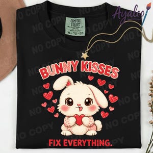 Könnte beinhalten: Schwarzes Sweatshirt mit dem Text "BUNNY KISSES FIX EVERYTHING" und einem Cartoon-Hasen, der ein Herz hält, umgeben von roten Herzen. Das Sweatshirt hat ein Comfort Colors-Etikett.