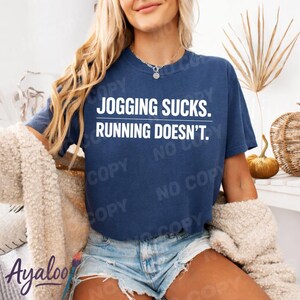 Puede incluir: Una camiseta azul marino con el texto "JOGGING SUCKS. RUNNING DOESN'T." en blanco. La camiseta se combina con pantalones cortos de mezclilla. La persona lleva una rebeca color crema.
