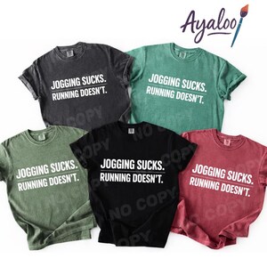 Puede incluir: Cinco camisetas de manga corta en varios colores: carb&oacute;n, verde, negro, verde oliva y granate. Cada camiseta muestra el texto "JOGGING SUCKS. RUNNING DOESN'T." en blanco. Las camisetas est&aacute;n dispuestas de forma informal.