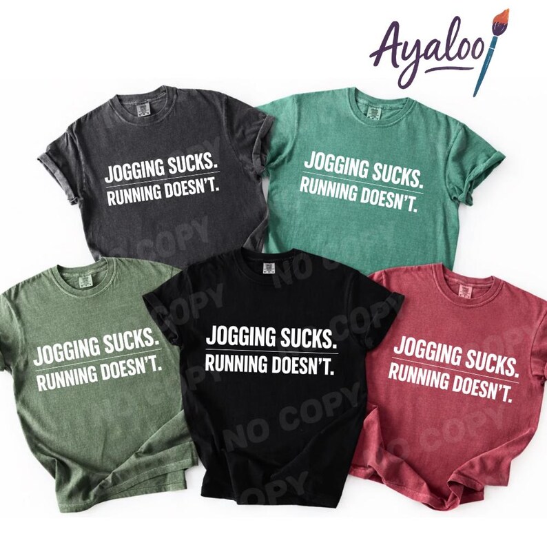 Puede incluir: Cinco camisetas de manga corta en varios colores: carb&oacute;n, verde, negro, verde oliva y granate. Cada camiseta muestra el texto "JOGGING SUCKS. RUNNING DOESN'T." en blanco. Las camisetas est&aacute;n dispuestas de forma informal.