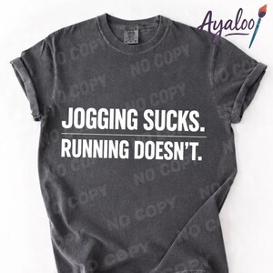 Puede incluir: Camiseta gris oscuro con mangas enrolladas. La camiseta presenta el texto blanco "JOGGING SUCKS. RUNNING DOESN'T." en negrita, con fuente sans-serif. La camiseta tiene un aspecto vintage y desgastado.