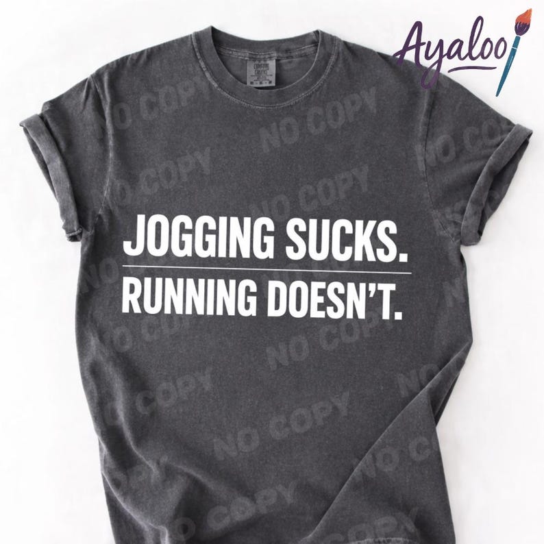 Puede incluir: Camiseta gris oscuro con mangas enrolladas. La camiseta presenta el texto blanco "JOGGING SUCKS. RUNNING DOESN'T." en negrita, con fuente sans-serif. La camiseta tiene un aspecto vintage y desgastado.