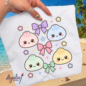 May include: A white t-shirt featuring a graphic print of four pastel-coloured, smiling dumpling characters in pink, blue, green, and yellow, interspersed with three bows in purple, pink, and green, along with small yellow and blue flower icons.