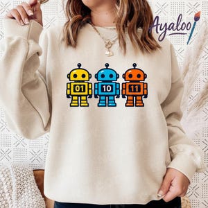 May include: Beige sweatshirt featuring three colorful robot graphics. The robots are yellow, blue, and orange, each with a different number: 01, 10, and 11. The robots have black outlines and are on a beige background.