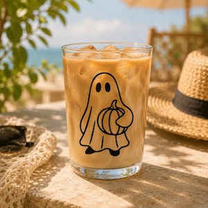 May include: A tall glass filled with iced coffee, featuring a black ghost and pumpkin design. The glass sits on a stone surface, with a straw hat and sunglasses in the background. The drink has a light brown color.