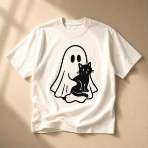 May include: Off-white t-shirt featuring a black graphic of a ghost and a black cat. The ghost has two oval eyes, and the cat is sitting. The shirt has short sleeves and a crew neck.