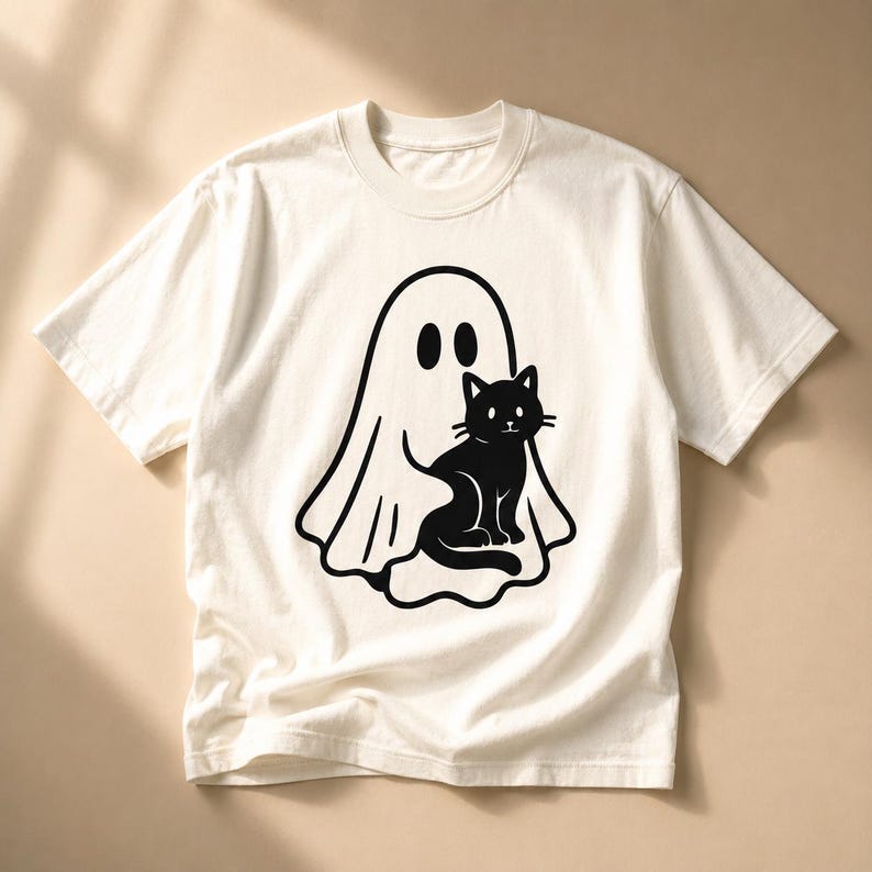 May include: Off-white t-shirt featuring a black graphic of a ghost and a black cat. The ghost has two oval eyes, and the cat is sitting. The shirt has short sleeves and a crew neck.