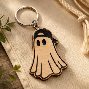 May include: A wooden ghost keychain with a black baseball cap. The ghost has two oval eyes and a wavy bottom edge. The keychain is attached to a silver metal ring.