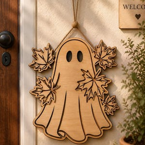 May include: Wooden ghost-shaped door hanger with maple leaf accents. The ghost has two oval eyes and is suspended by a jute rope. The door hanger is made of light-colored wood and is hanging on a beige wall.