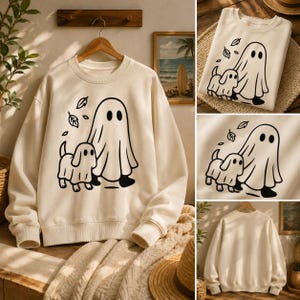 May include: Cream-colored sweatshirt featuring a black graphic of a ghost and a dog, with falling leaves. The design is centered on the front of the long-sleeved, crew-neck top. The sweatshirt is displayed on a wooden hanger.