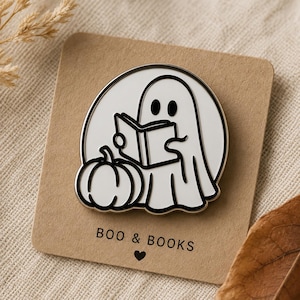 May include: A white enamel pin featuring a ghost reading a book next to a pumpkin. The pin has a black outline and the text "BOO & BOOKS" with a heart symbol. The pin is on a brown card.