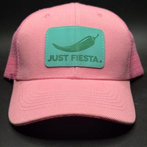 May include: Pink baseball cap with a mesh back and a curved brim. A teal patch on the front features a green chili pepper graphic and the text "JUST FIESTA."