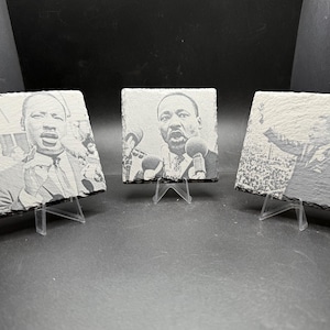 May include: Three slate coasters with engraved images of Martin Luther King Jr. Each coaster is displayed on a clear plastic stand. The images are in black and white, depicting MLK speaking at different events.