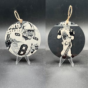 May include: Two round, black and white ornaments featuring football players. One ornament has the text "Sports Illustrated" and the number 8. The other ornament shows a player in action. Both have twine hangers.