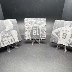May include: Three slate coasters featuring etched images of basketball players. Each coaster displays a different player in a jersey with a number and name. The coasters are displayed on clear plastic stands.
