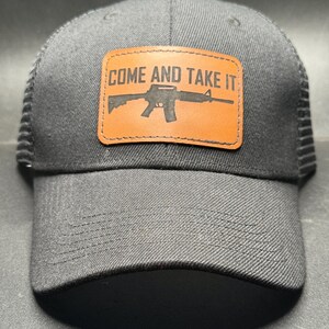 May include: A dark gray baseball cap with a mesh back and a curved brim. The cap features a rectangular, brown leather patch with the words "COME AND TAKE IT" and a rifle silhouette.