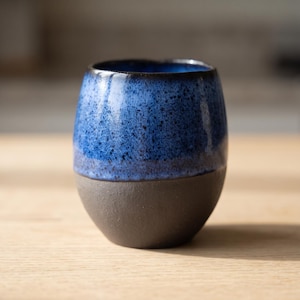 Peut inclure: Une tasse en céramique arrondie au design bicolore. La partie supérieure est dotée d'un émail bleu vif moucheté, tandis que la partie inférieure présente une base gris anthracite mat avec un aspect texturé et non émaillé.