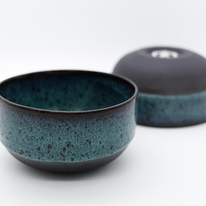 Peut inclure: Un ensemble de bol et de couvercle en céramique. Le bol a un intérieur bleu-vert foncé et un extérieur texturé bleu-vert et noir. Le couvercle est noir avec une bande bleu-vert à la base. L'ensemble est sur fond blanc.
