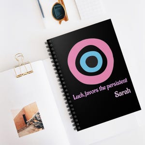 May include: Black spiral-bound notebook featuring a pink and blue target design. The text "Luck favors the persistent Sarah" is printed in light purple. The notebook is open on a white surface.
