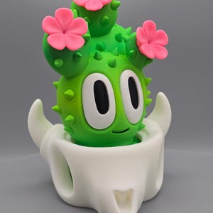 May include: A whimsical green cactus figurine with pink flower accents and large white eyes, set in a white skull-shaped planter. The cactus has a smiling face and is covered in small green spikes.