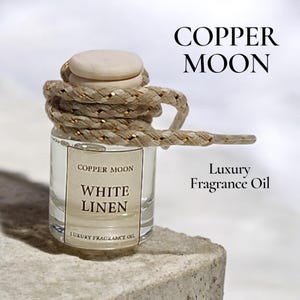 May include: A small glass bottle of "White Linen" luxury fragrance oil, with a wooden cap and a decorative rope. The bottle is labeled "Copper Moon". The rope is wrapped around the bottle and has gold accents.