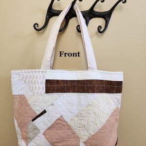 May include: A quilted tote bag with a patchwork design in shades of cream, tan, and brown. The bag features a geometric pattern with a brown rectangular accent and cream-colored handles. The word "Front" is printed on the bag.