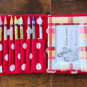May include: A red fabric crayon and photo album case with white polka dots. The case has a handle and a snap closure. It holds six crayons and a photo album with a plaid frame. The crayons are various colors.
