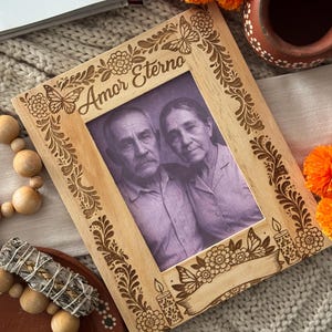 May include: A wooden picture frame with the words "Amor Eterno" engraved on it, featuring a black and white photo of an elderly couple. The frame is decorated with floral and butterfly designs.