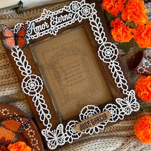 May include: A dark wood picture frame featuring white laser-cut floral and butterfly motifs. The top displays the text Amor Eterno, while a small wooden plaque at the bottom reads JOSE GARCIA. The center contains a card with Spanish text.