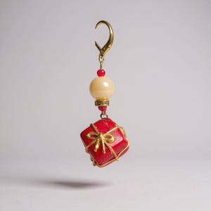 Peut inclure: Boucle d'oreille unique avec un charm en forme de boîte cadeau rouge, orné d'un nœud doré et de détails en fil d'or. La boucle d'oreille comprend une perle crème, une perle rouge et un fermoir doré. L'ensemble est suspendu sur fond blanc.