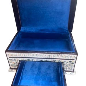 May include: A dark wood jewelry box with a hinged lid and a pull-out drawer. The interior is lined with bright blue velvet. The exterior features intricate geometric mother-of-pearl inlay patterns along the front and sides, with a small white knob.