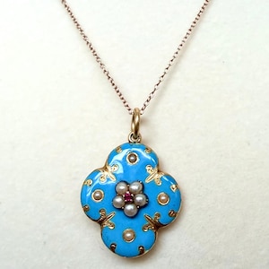 May include: A gold pendant on a delicate chain featuring a quatrefoil shape with bright blue enamel, gold filigree accents, and a central flower motif composed of small white seed pearls surrounding a single tiny red gemstone.