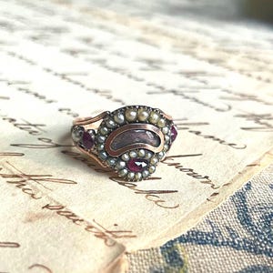 Op de afbeelding: Een vintage ring met een gouden band en een centraal ontwerp met een donkere, ovale steen, omringd door kleine parels en rode edelstenen. De ring ligt op crèmekleurig papier met handschrift.